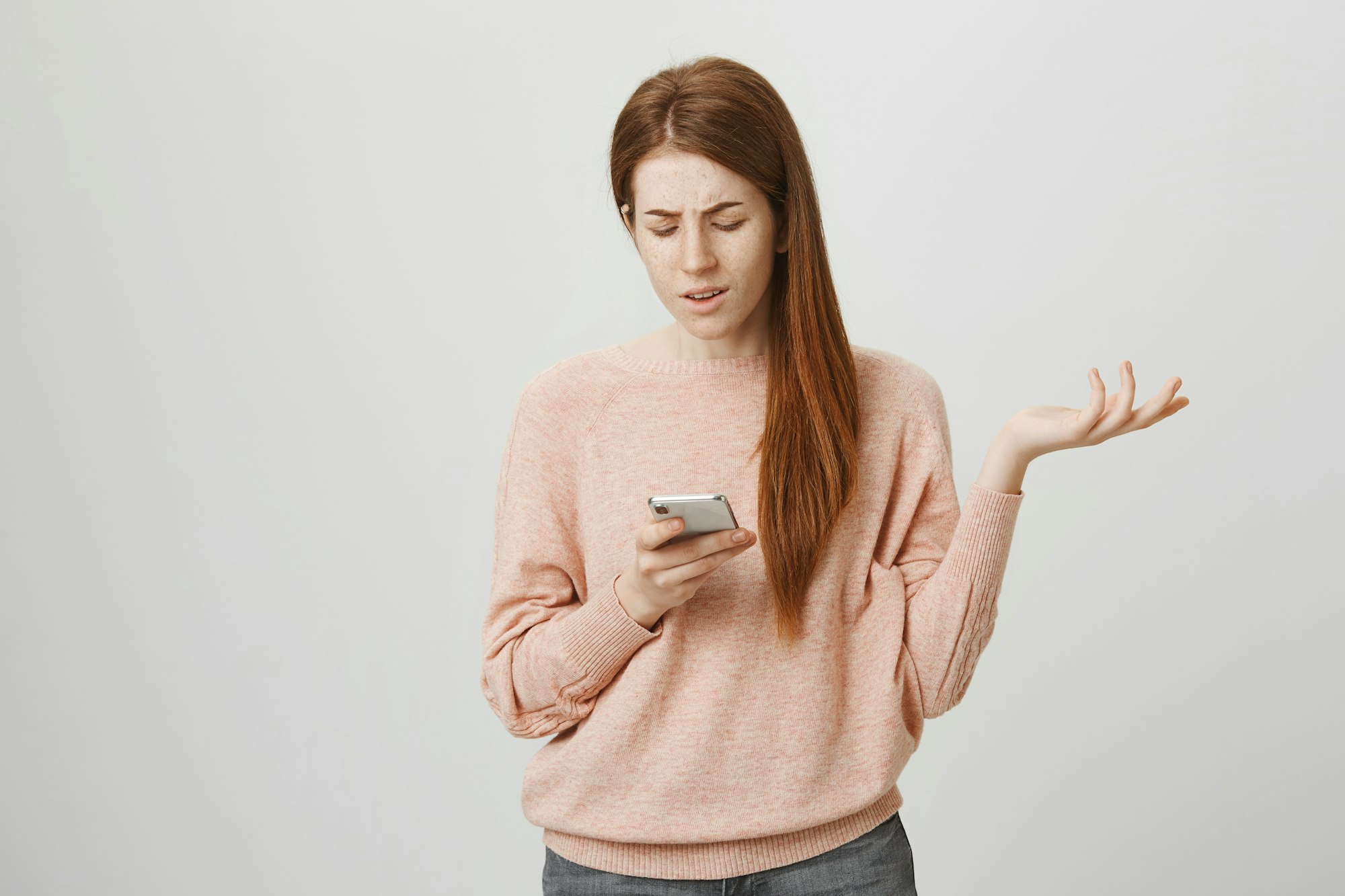 Upset or irritated ginger girl gesturing while looking at smartphone screen, being clueless or